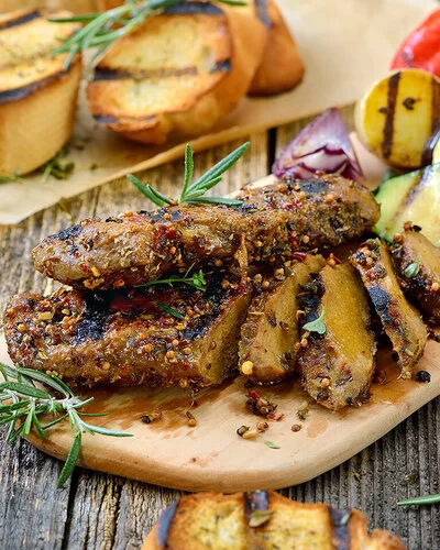 Auf einem Holzbrett liegen in Scheiben geschnittene Seitan-Steaks mit gegrillten Zucchini, Möhren, Zwiebeln und Maiskolben. Im Hintergrund sind gegrillte Baguettescheiben zu sehen.