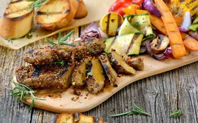 Auf einem Holzbrett liegen in Scheiben geschnittene Seitan-Steaks mit gegrillten Zucchini, Möhren, Zwiebeln und Maiskolben. Im Hintergrund sind gegrillte Baguettescheiben zu sehen.