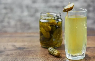 Auf einem Holztisch steht ein Glas mit hellgelbem Gurkenwasser. Es ist mit einer aufgespießten Essiggurkenscheibe garniert. Im Hintergrund ergänzen ein Glas eingelegter Gurken und eine einzelne Essiggurke die Szene.