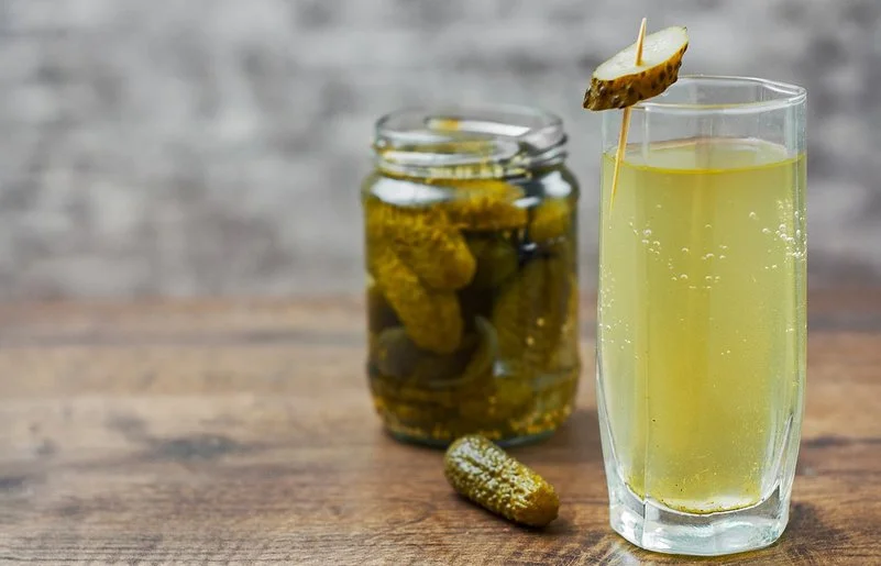 Auf einem Holztisch steht ein Glas mit hellgelbem Gurkenwasser. Es ist mit einer aufgespießten Essiggurkenscheibe garniert. Im Hintergrund ergänzen ein Glas eingelegter Gurken und eine einzelne Essiggurke die Szene.