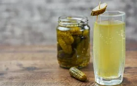 Auf einem Holztisch steht ein Glas mit hellgelbem Gurkenwasser. Es ist mit einer aufgespießten Essiggurkenscheibe garniert. Im Hintergrund ergänzen ein Glas eingelegter Gurken und eine einzelne Essiggurke die Szene.