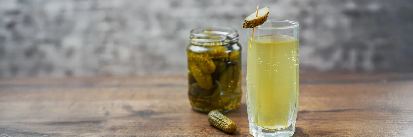 Auf einem Holztisch steht ein Glas mit hellgelbem Gurkenwasser. Es ist mit einer aufgespießten Essiggurkenscheibe garniert. Im Hintergrund ergänzen ein Glas eingelegter Gurken und eine einzelne Essiggurke die Szene.
