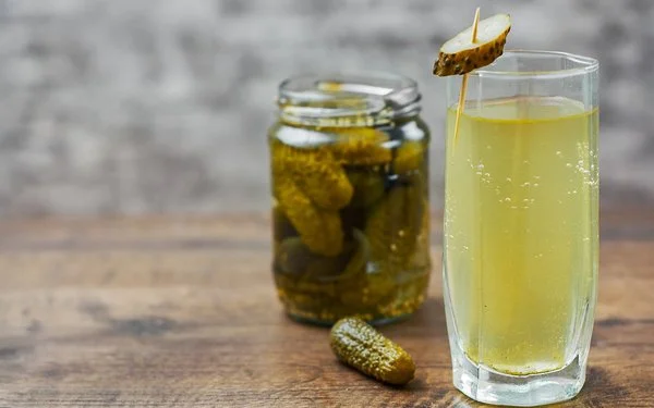 Auf einem Holztisch steht ein Glas mit hellgelbem Gurkenwasser. Es ist mit einer aufgespießten Essiggurkenscheibe garniert. Im Hintergrund ergänzen ein Glas eingelegter Gurken und eine einzelne Essiggurke die Szene.
