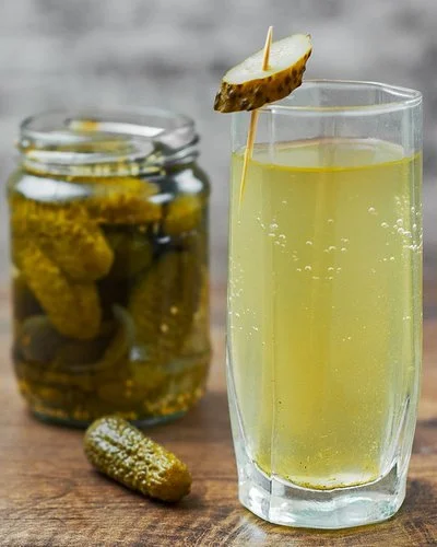 Auf einem Holztisch steht ein Glas mit hellgelbem Gurkenwasser. Es ist mit einer aufgespießten Essiggurkenscheibe garniert. Im Hintergrund ergänzen ein Glas eingelegter Gurken und eine einzelne Essiggurke die Szene.