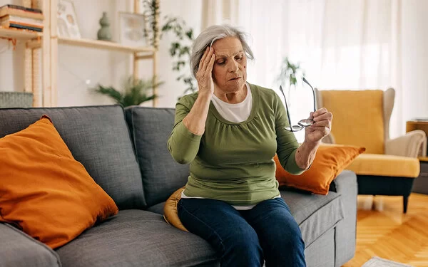 Eine ältere Frau fasst sich leicht benommen und mit abgesetzter Brille an den Kopf.