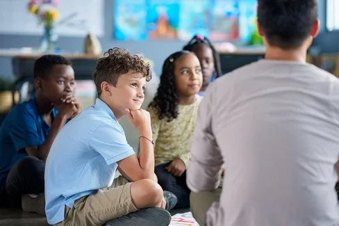 Ein Gruppe von Kindern sitzt in einem Kreis auf dem Boden. Sie blicken auf eine erwachsene Person, die vor ihnen sitzt.