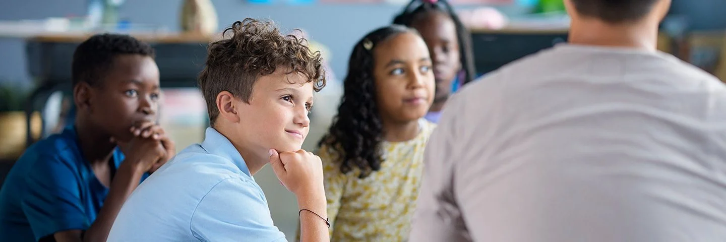Ein Gruppe von Kindern sitzt in einem Kreis auf dem Boden. Sie blicken auf eine erwachsene Person, die vor ihnen sitzt.