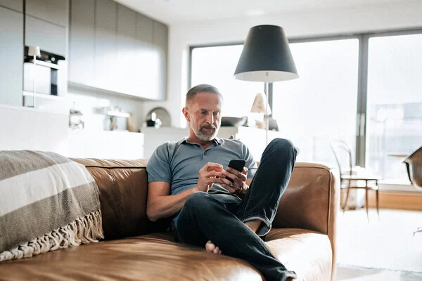 Ein Mann sitzt auf einer Couch. Er blickt auf das Smartphone, das er in der Hand hält.