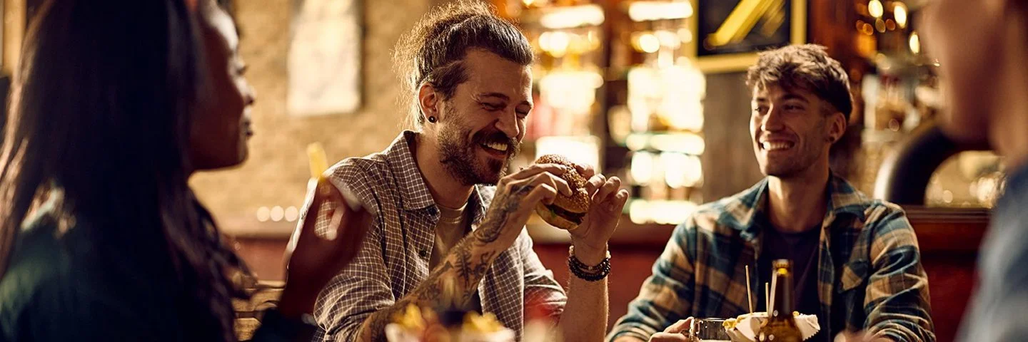 Ein lachender Mann mit einem Burger in der Hand sitzt abends mit Freunden an einem Tisch in einem Restaurant.
