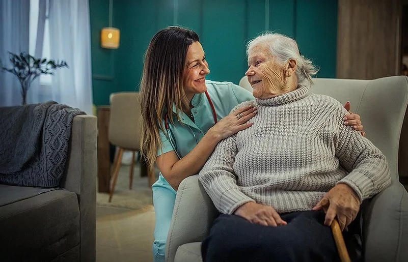 Eine alte Frau sitzt in einem Sessel. Neben ihr kniet eine Pflegekraft und legt ihre Hände auf die Schultern der Frau. Sie sehen sich in die Augen und lächeln.