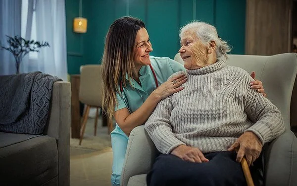 Eine alte Frau sitzt in einem Sessel. Neben ihr kniet eine Pflegekraft und legt ihre Hände auf die Schultern der Frau. Sie sehen sich in die Augen und lächeln.