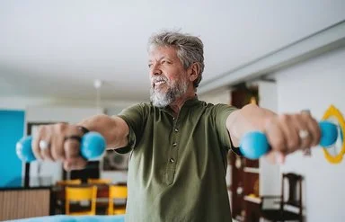Ein älterer Mann mit grauen Haaren und grauem Bart hebt zwei türkise Hanteln und schaut dabei in die Ferne.
