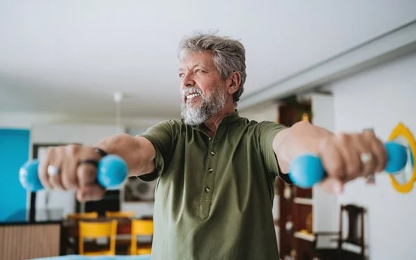 Ein älterer Mann mit grauen Haaren und grauem Bart hebt zwei türkise Hanteln und schaut dabei in die Ferne.