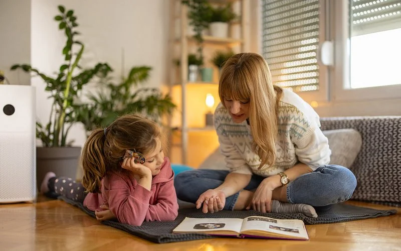 Frau und Kind liegen auf einem Teppich am Boden und schauen gemeinsam in ein aufgeschlagenes Buch; gemütliche Wohnumgebung im Hintergrund.
