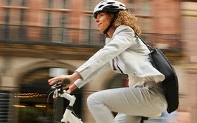 Eine Frau fährt mit Fahrradhelm und in einem weißen Blazer dynamisch auf ihrem Rad zur Arbeit.