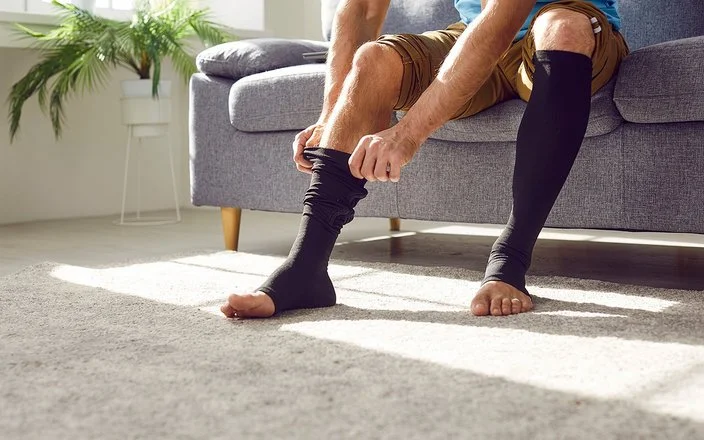 Ein Mann sitzt auf einem Sofa und zieht sich lange schwarze Thrombosestrümpfe an. Der Oberkörper ist im Bildausschnitt nicht erfasst.