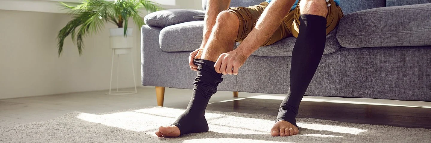 Ein Mann sitzt auf einem Sofa und zieht sich lange schwarze Thrombosestrümpfe an. Der Oberkörper ist im Bildausschnitt nicht erfasst.