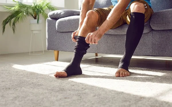 Ein Mann sitzt auf einem Sofa und zieht sich lange schwarze Thrombosestrümpfe an. Der Oberkörper ist im Bildausschnitt nicht erfasst.