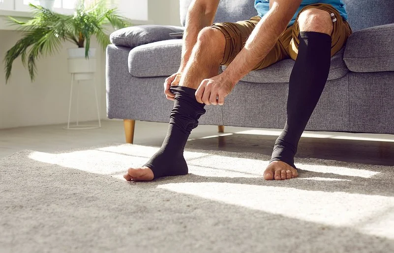 Ein Mann sitzt auf einem Sofa und zieht sich lange schwarze Thrombosestrümpfe an. Der Oberkörper ist im Bildausschnitt nicht erfasst.