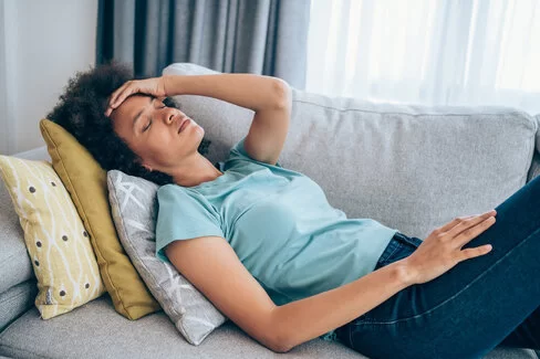 Eine Frau mit Post-COVID-Symptomen liegt auf einem Sofa und hält sich mit schmerzverzerrtem Gesicht die Stirn.