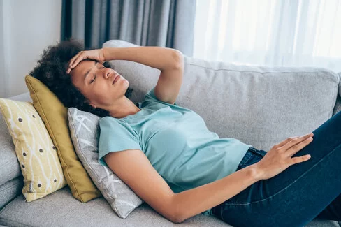 Eine Frau mit Post-COVID-Symptomen liegt auf einem Sofa und hält sich mit schmerzverzerrtem Gesicht die Stirn.