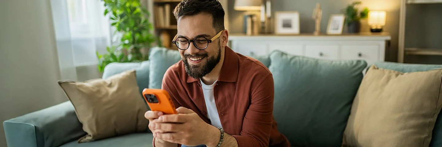 Ein Mann mit Brille und Bart sitzt lächelnd auf einem Sofa und schaut auf ein orangefarbenes Smartphone.
