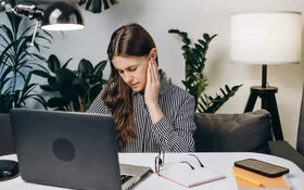 Eine junge Frau sitzt vor ihrem Laptop am Schreibtisch und fasst sich ans Ohr.