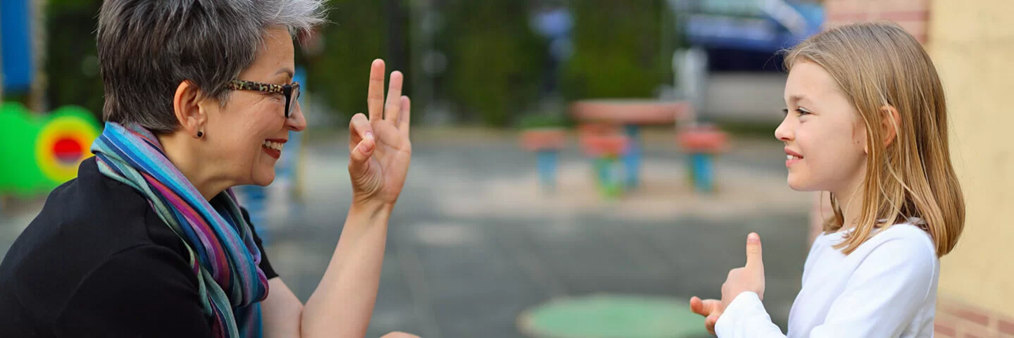 Mutter und Tochter sind auf einem Spielplatz und sitzen in einem kleinen Karussell, sie unterhalten sich in Gebärdensprache miteinander.