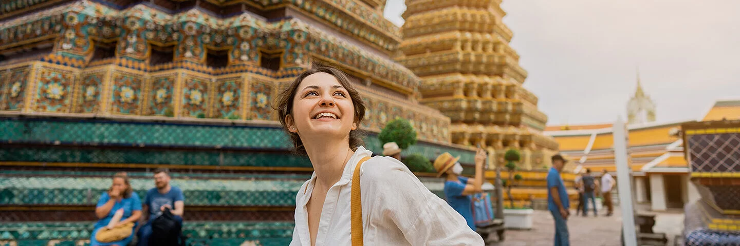 Eine Frau mit Rucksack steht lächelnd vor dem kunstvoll verzierten Tempel Wat Pho in Bangkok und schaut nach oben.