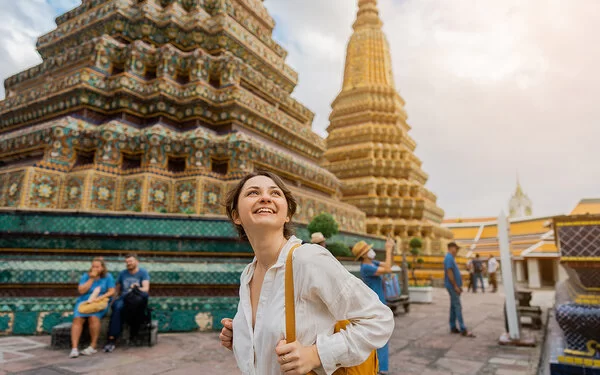 Eine Frau mit Rucksack steht lächelnd vor dem kunstvoll verzierten Tempel Wat Pho in Bangkok und schaut nach oben.