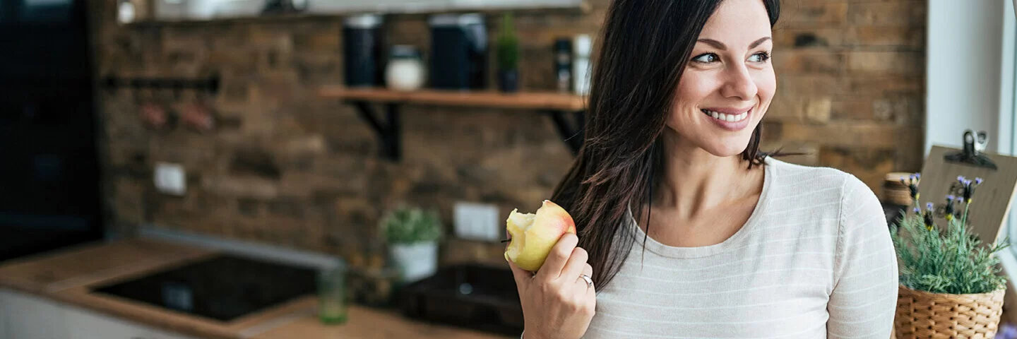 Junge Frau steht am Küchenfenster und isst einen Apfel, um ihr Immunsystem zu stärken.