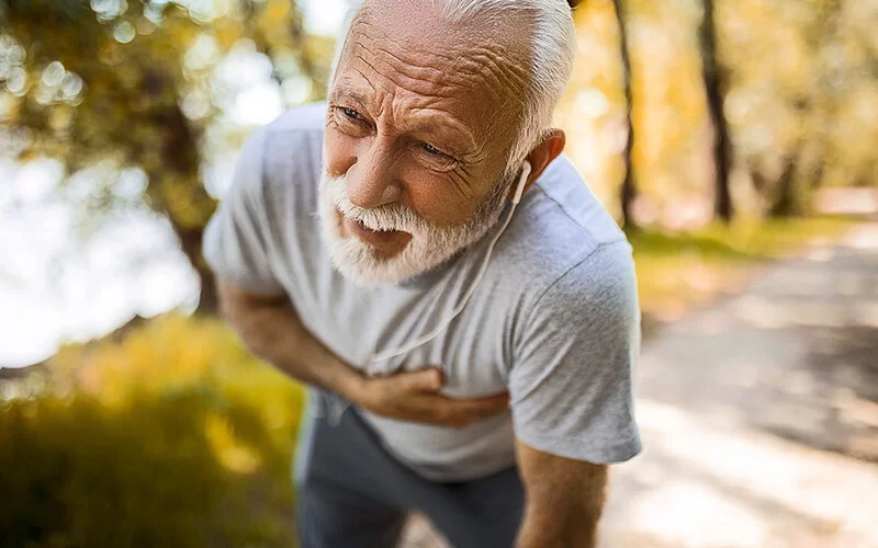 Ein älterer Mann, der im Park joggt, geht leicht in die Knie und fasst sich mit einer Hand auf das Herz.