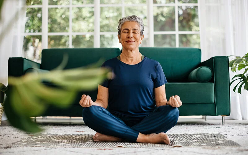 Eine ältere Frau mit kurzen grauen Haaren sitzt im Schneidersitz auf einer Yogamatte im Wohnzimmer und meditiert.