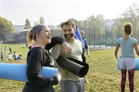 Zwei Männer und zwei Frauen stehen mit Yoga-Matten unter den Armen auf einer Wiese und freuen sich.