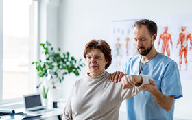 Eine ältere Frau sitzt auf einer Behandlungsliege mit dem Rücken zu einem Arzt, der ihre linke Schulter untersucht.