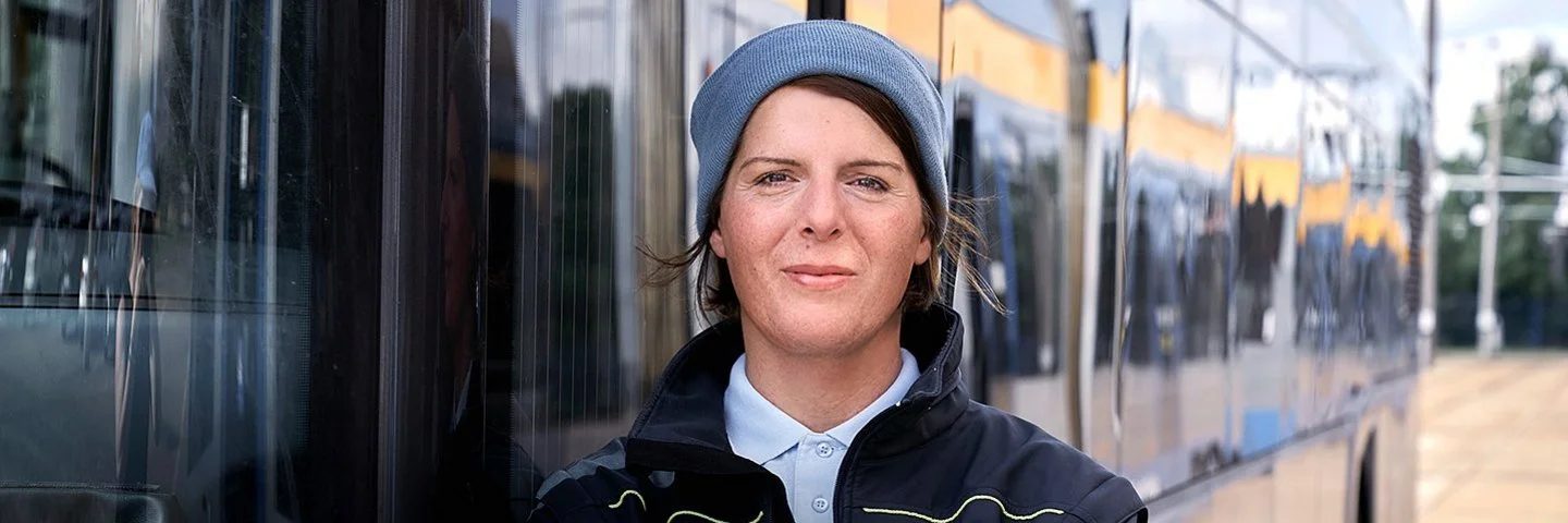 Die Busfahrerin Angela Rossberg steht mit vor dem Oberkörper verschränkten Armen vor einem Bus und schaut selbstbewusst in die Kamera.