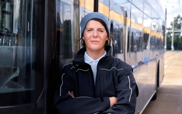 Die Busfahrerin Angela Rossberg steht mit vor dem Oberkörper verschränkten Armen vor einem Bus und schaut selbstbewusst in die Kamera.