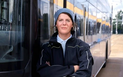 Die Busfahrerin Angela Rossberg steht mit vor dem Oberkörper verschränkten Armen vor einem Bus und schaut selbstbewusst in die Kamera.