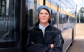 Die Busfahrerin Angela Rossberg steht mit vor dem Oberkörper verschränkten Armen vor einem Bus und schaut selbstbewusst in die Kamera.