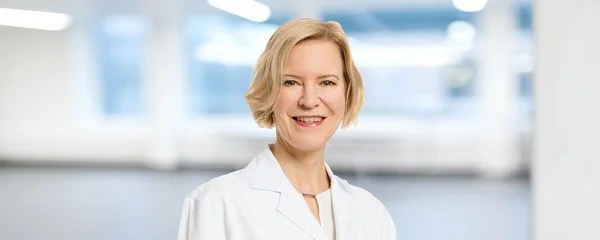 Die Professorin Dr. Petra Stute trägt einen weißen Laborkittel. Der Hintergrund ist unscharf und zeigt einen hellen, modernen Innenraum mit großen Fenstern und Deckenbeleuchtung.