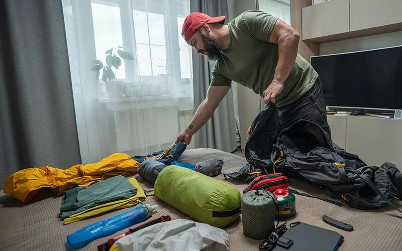 Ein Mann packt in seiner Wohnung seinen Rucksack für eine Trekking-Tour. Auf dem Bett vor ihm liegen wichtige Trekking-Utensilien ausgebreitet, darunter ein Zelt, ein Schlafsack, Funktionskleidung und eine Gasflasche.