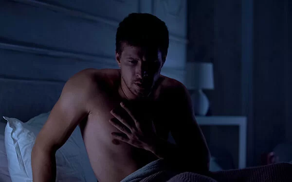 Mann sitzt nachts aufrecht im Bett und hält seine linke Hand auf der Brust.