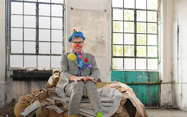 Ein Clown sitzt lachend auf Säcken in einer alten Lagerhalle.