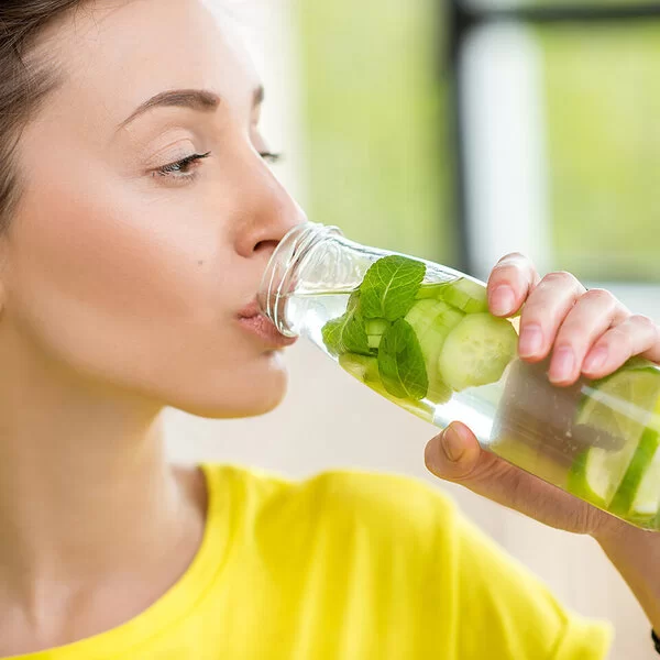 Eine junge Frau mit zurückgebundenen braunen Haaren trinkt aus einer Glasflasche Wasser, das mit Minzblättern sowie Gurken- und Limettenscheiben aromatisiert ist.