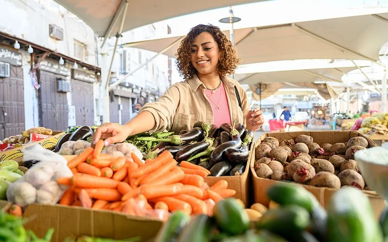 Eine junge Frau mit lockigen Haaren greift an einem Marktstand lachend nach einer Karotte.
