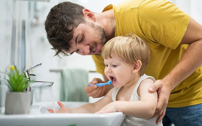 Ein ungefähr zweijähriger Junge und sein Vater im Bad. Der Junge steht auf einem Hocker am Waschbecken, der Vater beugt sich von hinten über ihn und hilft beim Zähneputzen.