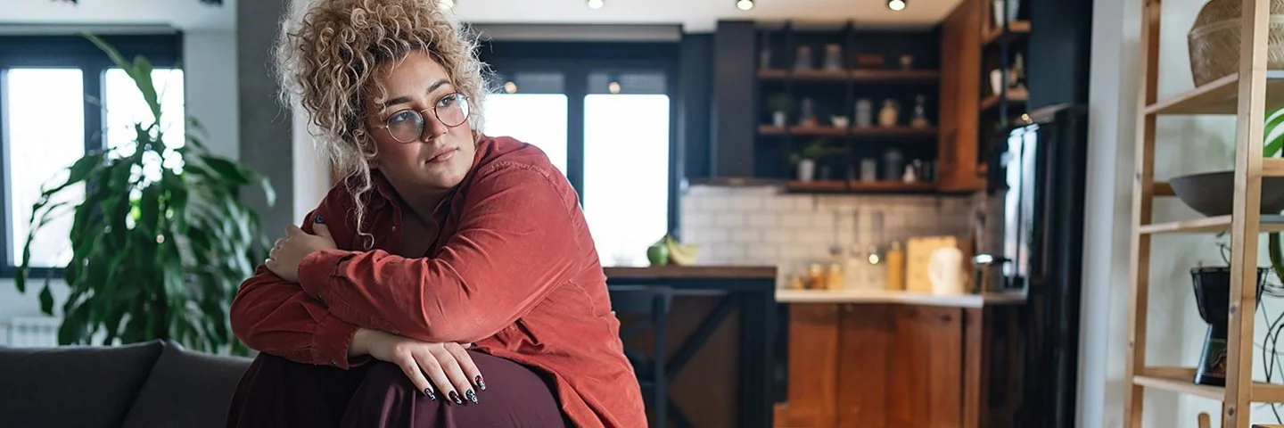 Eine mehrgewichtige Frau sitzt im Wohnzimmer auf dem Sofa, vor ihr steht eine Schachtel mit Pizza.