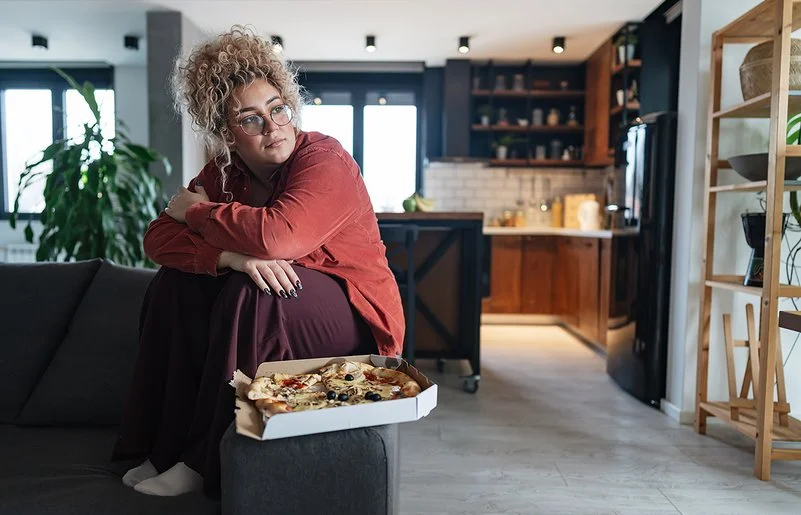 Eine mehrgewichtige Frau sitzt im Wohnzimmer auf dem Sofa, vor ihr steht eine Schachtel mit Pizza.