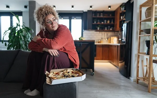 Eine mehrgewichtige Frau sitzt im Wohnzimmer auf dem Sofa, vor ihr steht eine Schachtel mit Pizza.