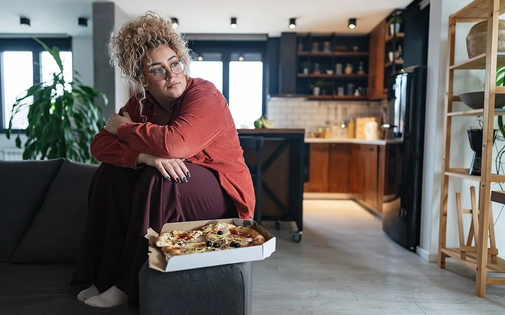Eine mehrgewichtige Frau sitzt im Wohnzimmer auf dem Sofa, vor ihr steht eine Schachtel mit Pizza.